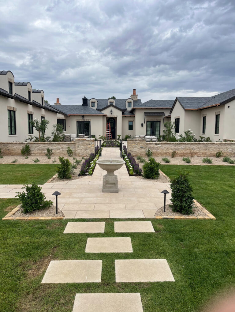 Backyard basketball court with green and blue painted surface, bordered by stone walls and young plants, set against a backdrop of palm trees and greenery.
