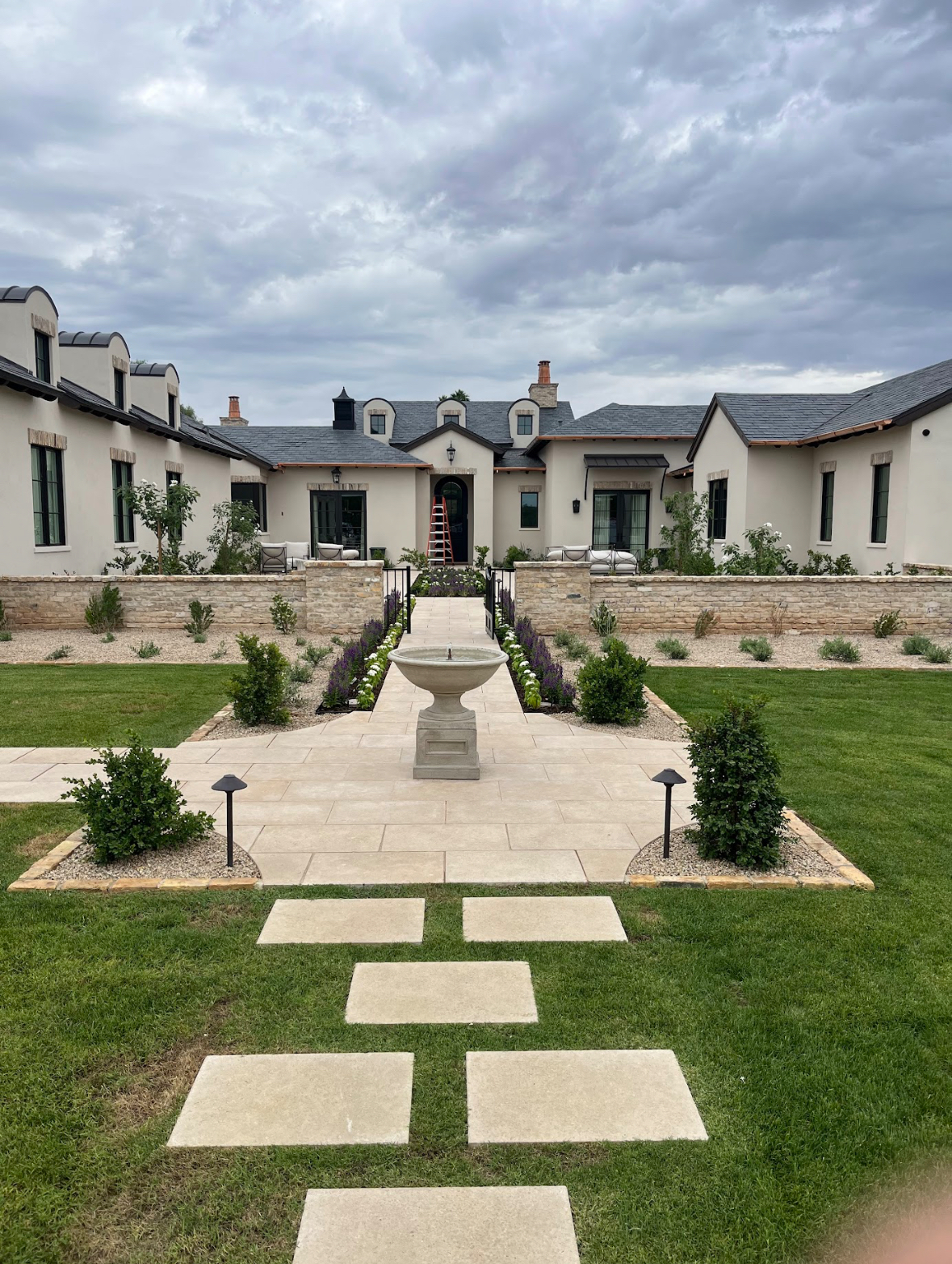 Backyard basketball court with green and blue painted surface, bordered by stone walls and young plants, set against a backdrop of palm trees and greenery.
