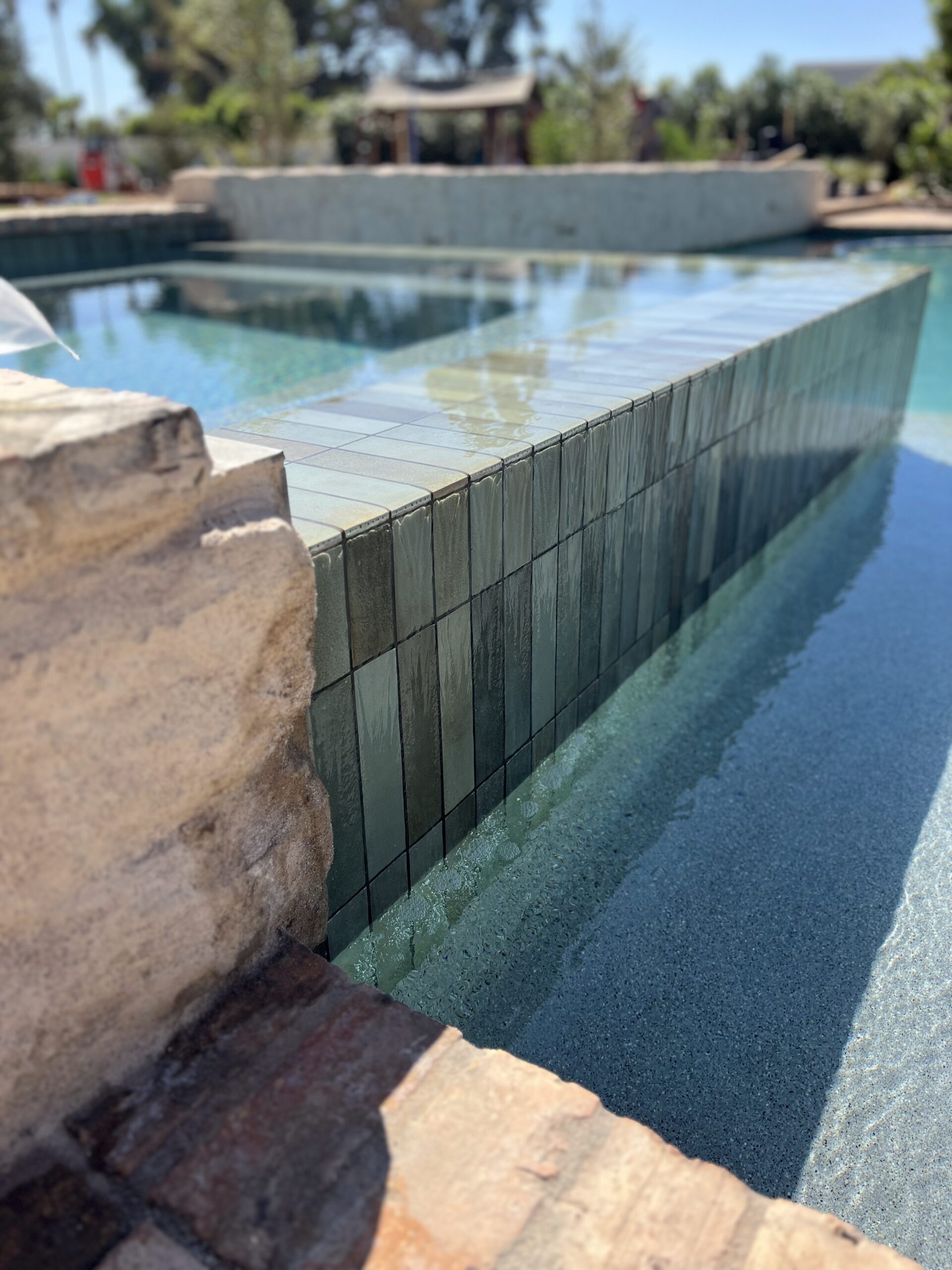 Close-up of a pool with raised tiled wall feature, clear water, and stone edging in an outdoor setting.