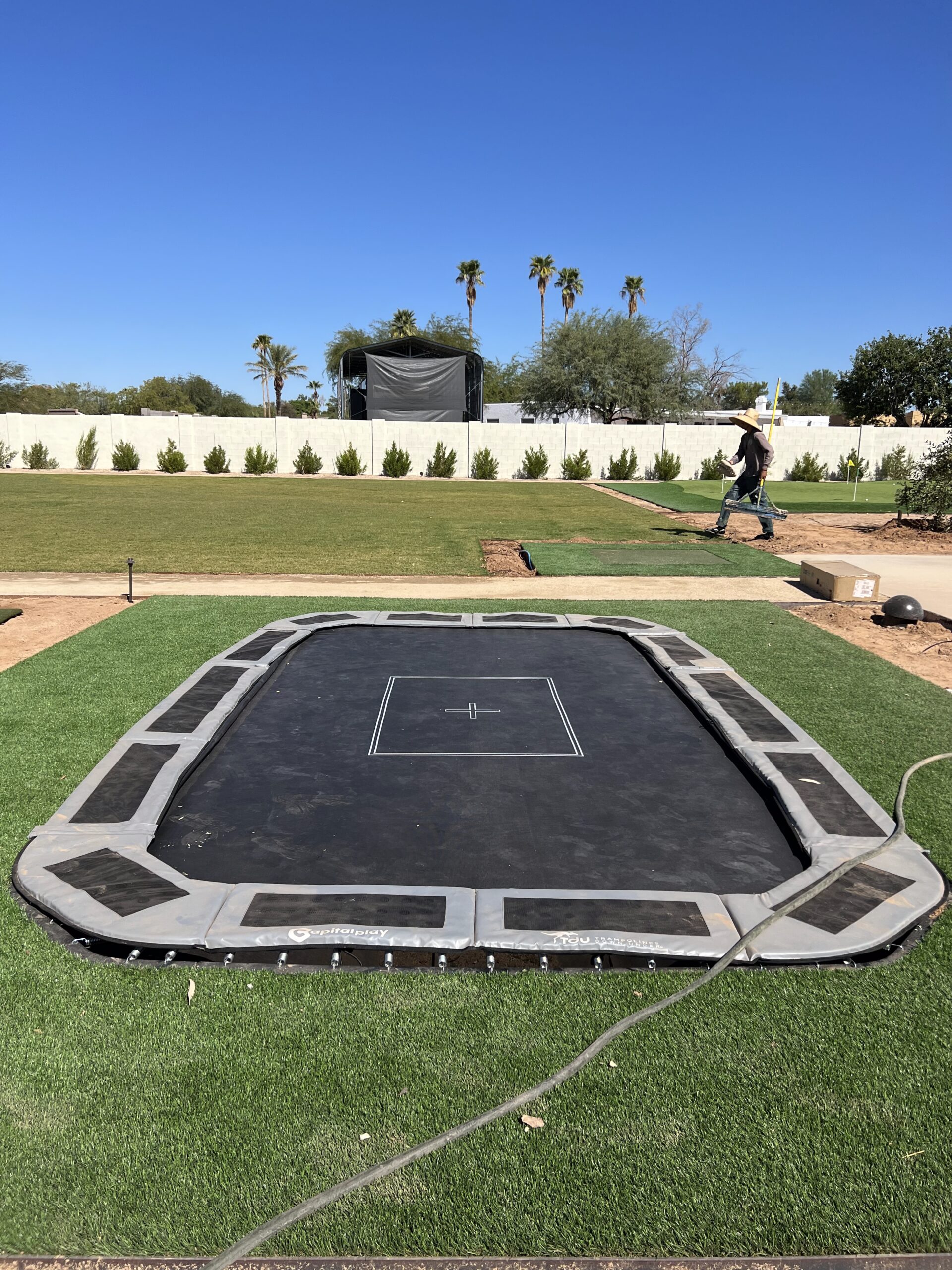 In-ground rectangular trampoline set with a landscaped yard, palm trees, and a person walking in the background